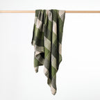 A green and beige plaid woven throw blanket draped over a light wood hanging rod against a plain white background.