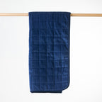 A blue quilted throw with a plush velvet texture, hanging from a light wood dowel against a clean white background.