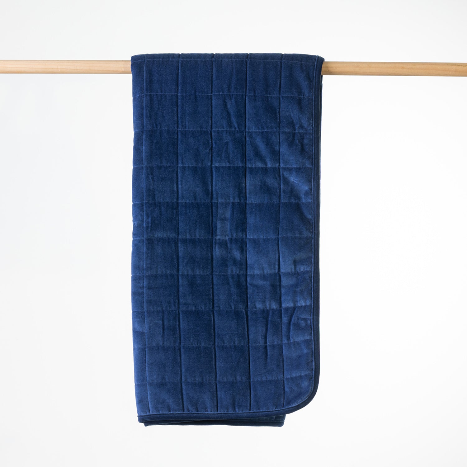 A blue quilted throw with a plush velvet texture, hanging from a light wood dowel against a clean white background.