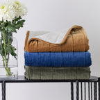 Three folded plush throws in rust, blue, and olive green are stacked on a black table, with a glass vase of white flowers on the left against a light grey background.