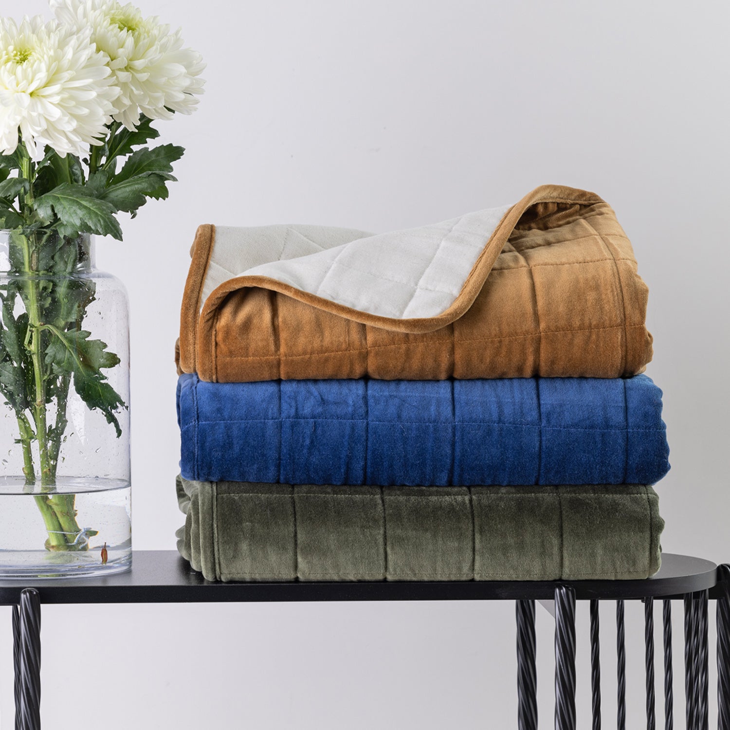 Three folded plush throws in rust, blue, and olive green are stacked on a black table, with a glass vase of white flowers on the left against a light grey background.