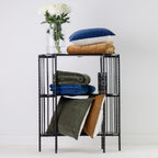 A black metal three-tier side table styled with folded rust, blue and olive green throws, cushions and a clear glass vase of white flowers, set against a white wall.