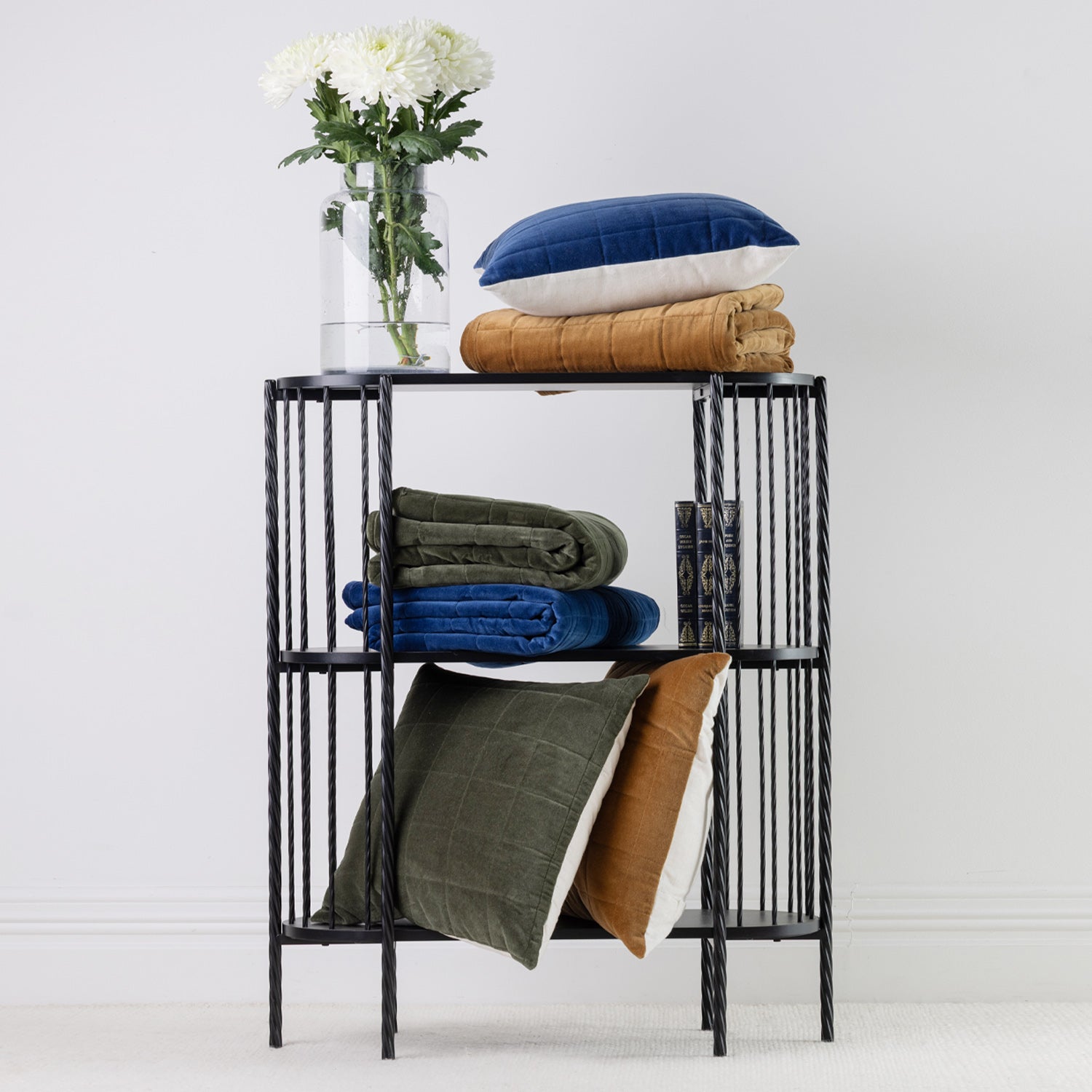 A black metal three-tier side table styled with folded rust, blue and olive green throws, cushions and a clear glass vase of white flowers, set against a white wall.