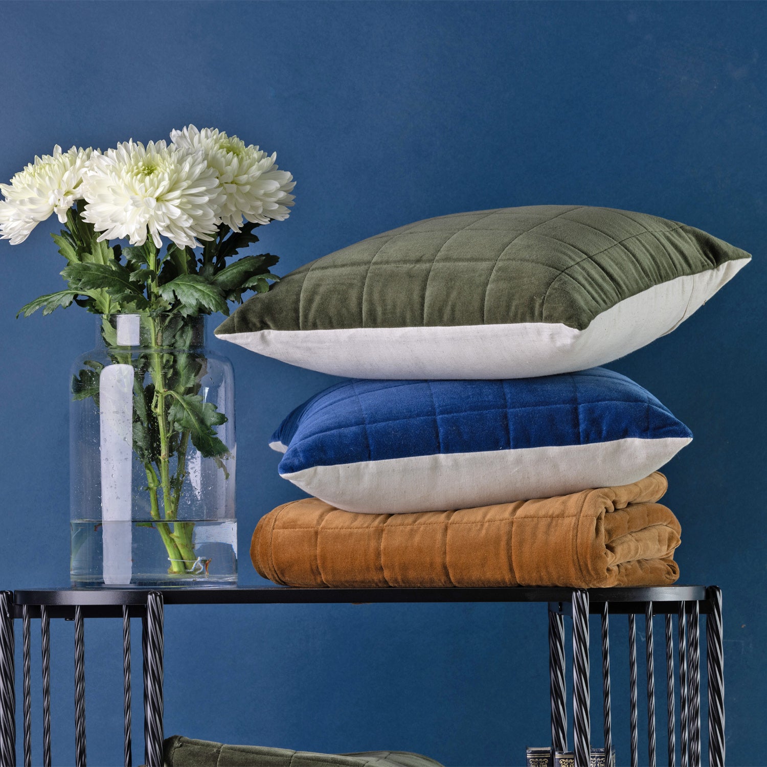 Quilted cushions & throw stacked on a black metal table against a dark blue wall. A clear glass vase with white flowers sits to the left.