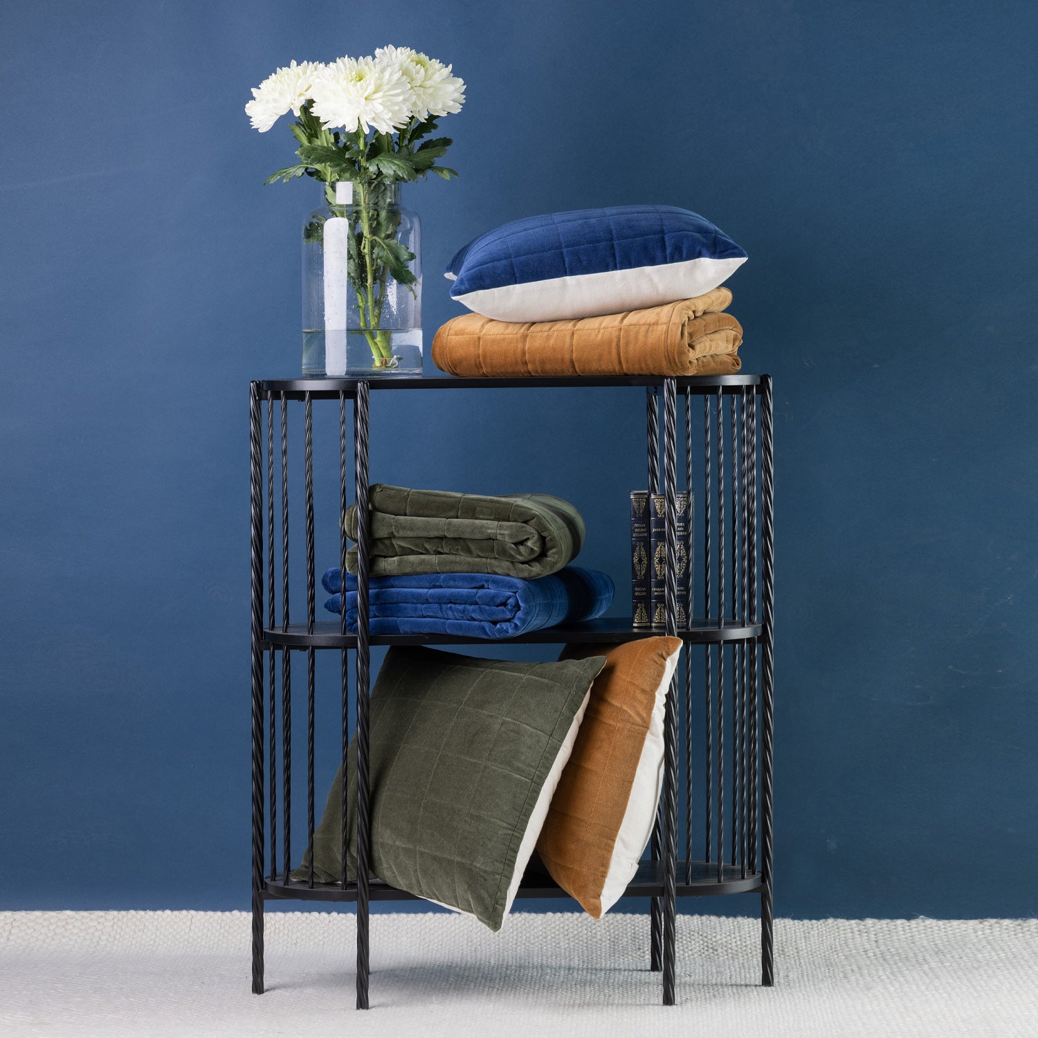 A black metal three-tier side table styled with folded rust, blue and olive green throws, cushions and a clear glass vase of white flowers, set against a dark blue wall.