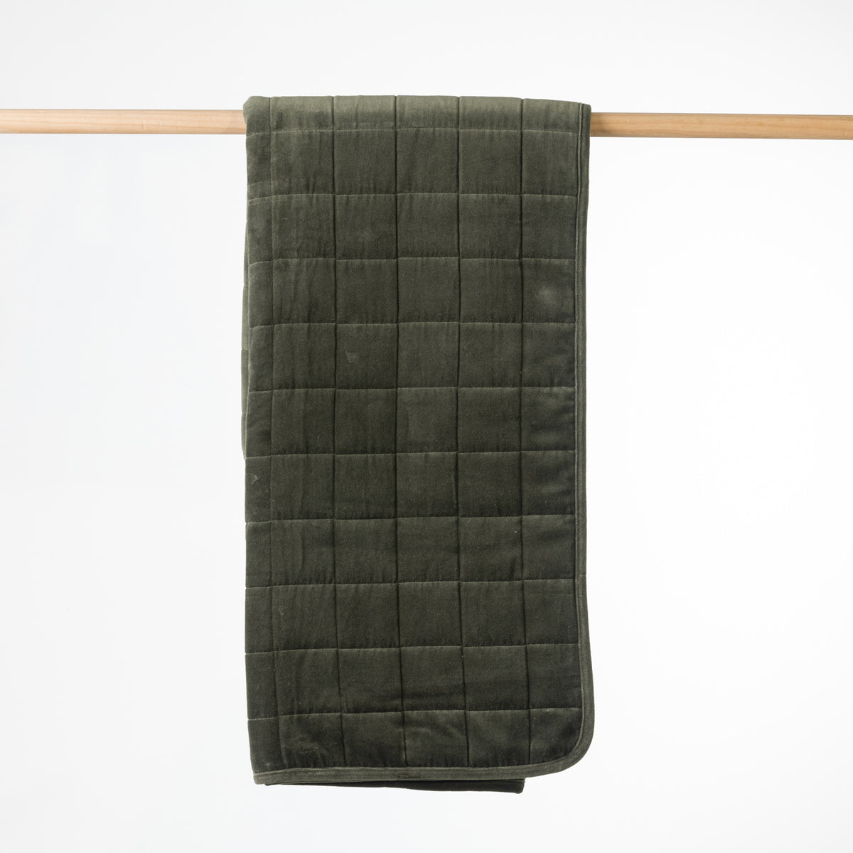 A olive quilted throw with a plush velvet texture, hanging from a light wood dowel against a clean white background.