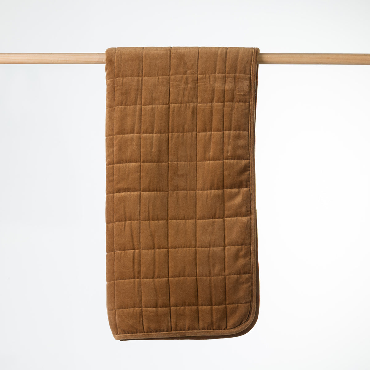 A rust quilted throw with a plush velvet texture, hanging from a light wood dowel against a clean white background.