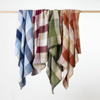Four woven throws with plaid patterns in blue, red, green, and rust hang from a light wood rod against a clean white background.