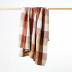 A textured waffle throw with a warm rust plaid pattern, hanging from a light wood rod against a clean white background.