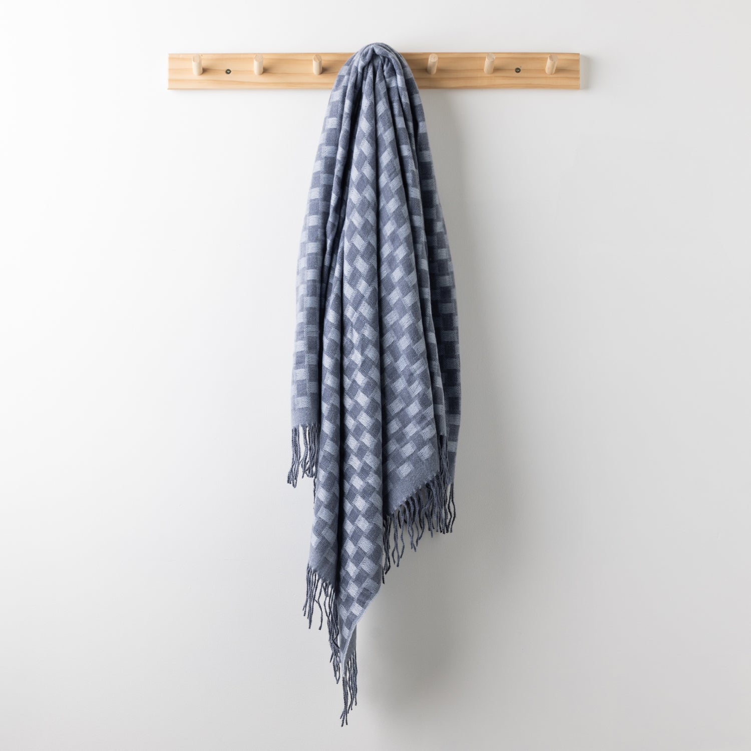 Home Co. Solane Check Throw in Blue featuring a blue check pattern and fringed edges, hanging from a wooden wall rack.