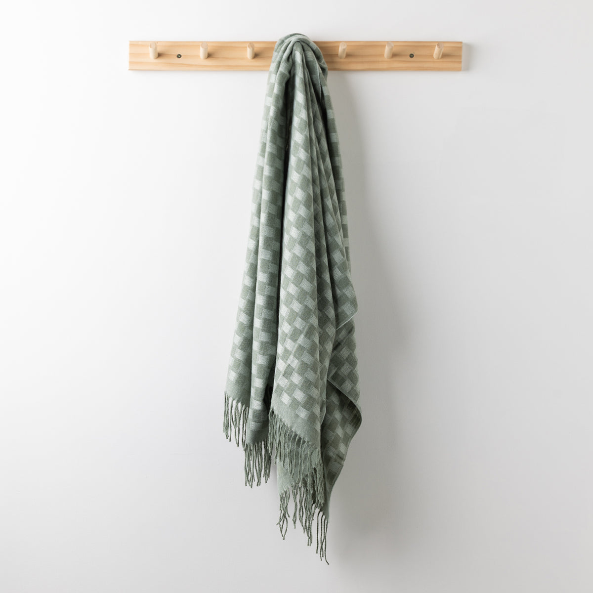Home Co. Solane Check Throw in Sage featuring a soft green check pattern and fringed edges, hanging from a wooden wall rack.