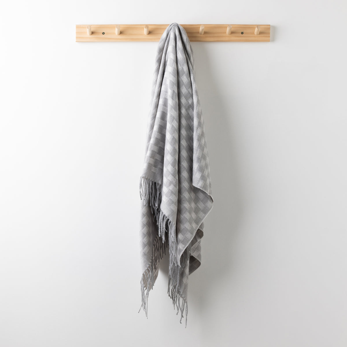 Home Co. Solane Check Throw in Silver featuring a light grey check pattern and fringed edges, hanging from a wooden wall rack.