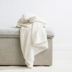 Almond: Home Co. Fraser Throw in soft almond, draped over a neutral upholstered ottoman in a light interior.