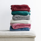 Home Co. Fraser Throws in wine, green, grey, almond, blush and blue, folded and stacked on a neutral upholstered ottoman against a white background.