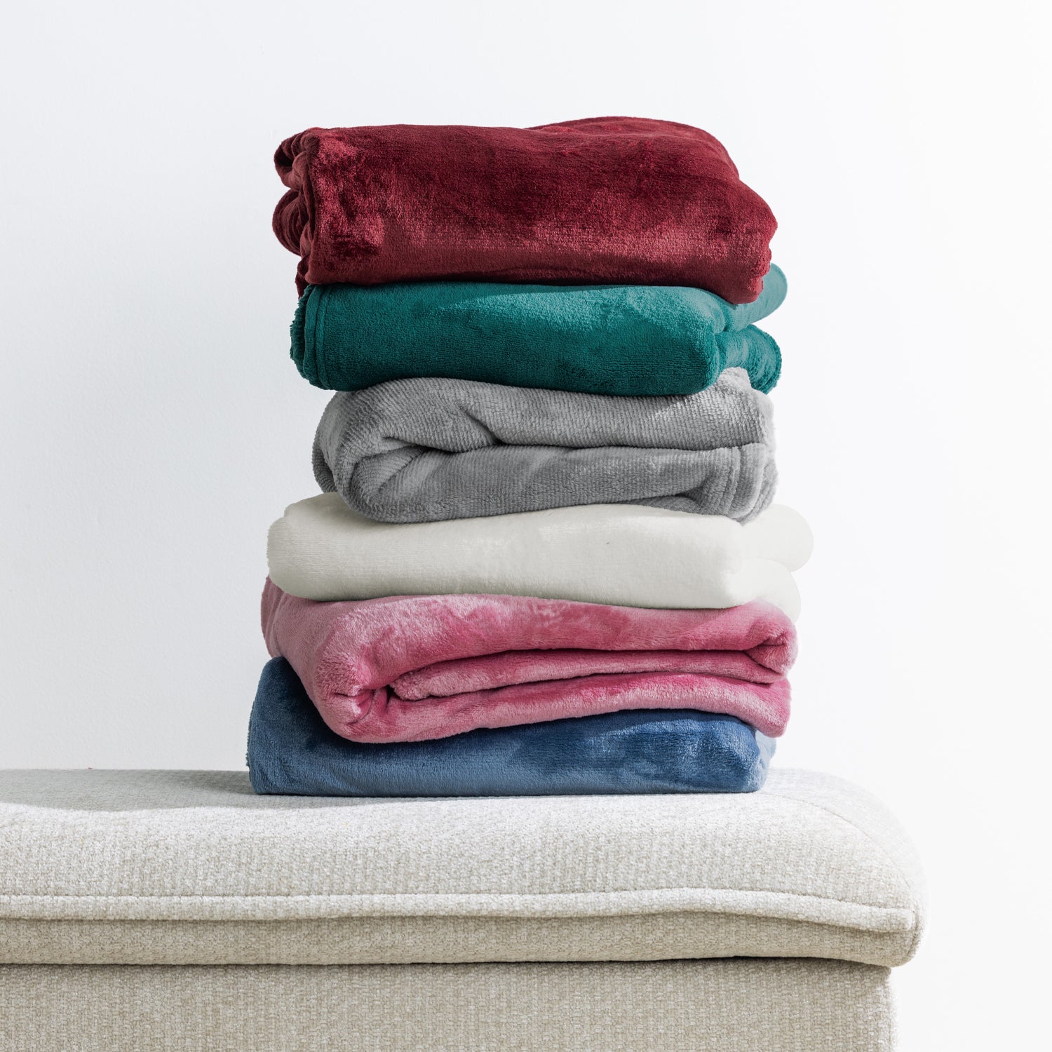 Home Co. Fraser Throws in wine, green, grey, almond, blush and blue, folded and stacked on a neutral upholstered ottoman against a white background.