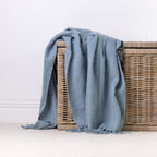 A blue faux mohair throw with fringe, draped over a wicker storage basket against a clean white wall and light carpet.