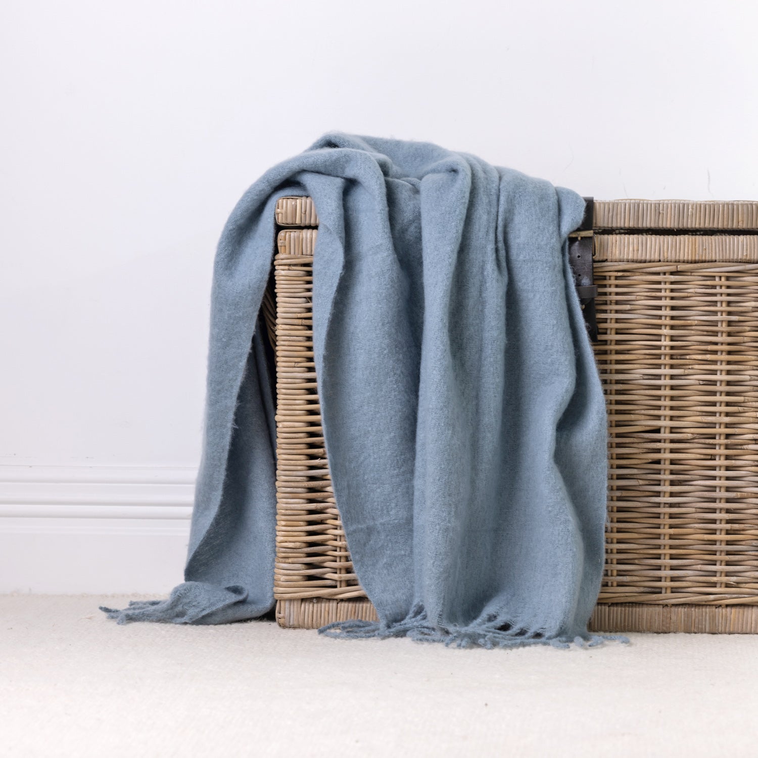 A blue faux mohair throw with fringe, draped over a wicker storage basket against a clean white wall and light carpet.
