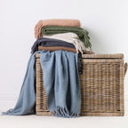 A stack of folded and draped faux mohair throws in blue, cocoa, green, natural, sand, and grey, arranged on and around a woven wicker storage basket against a plain white wall.