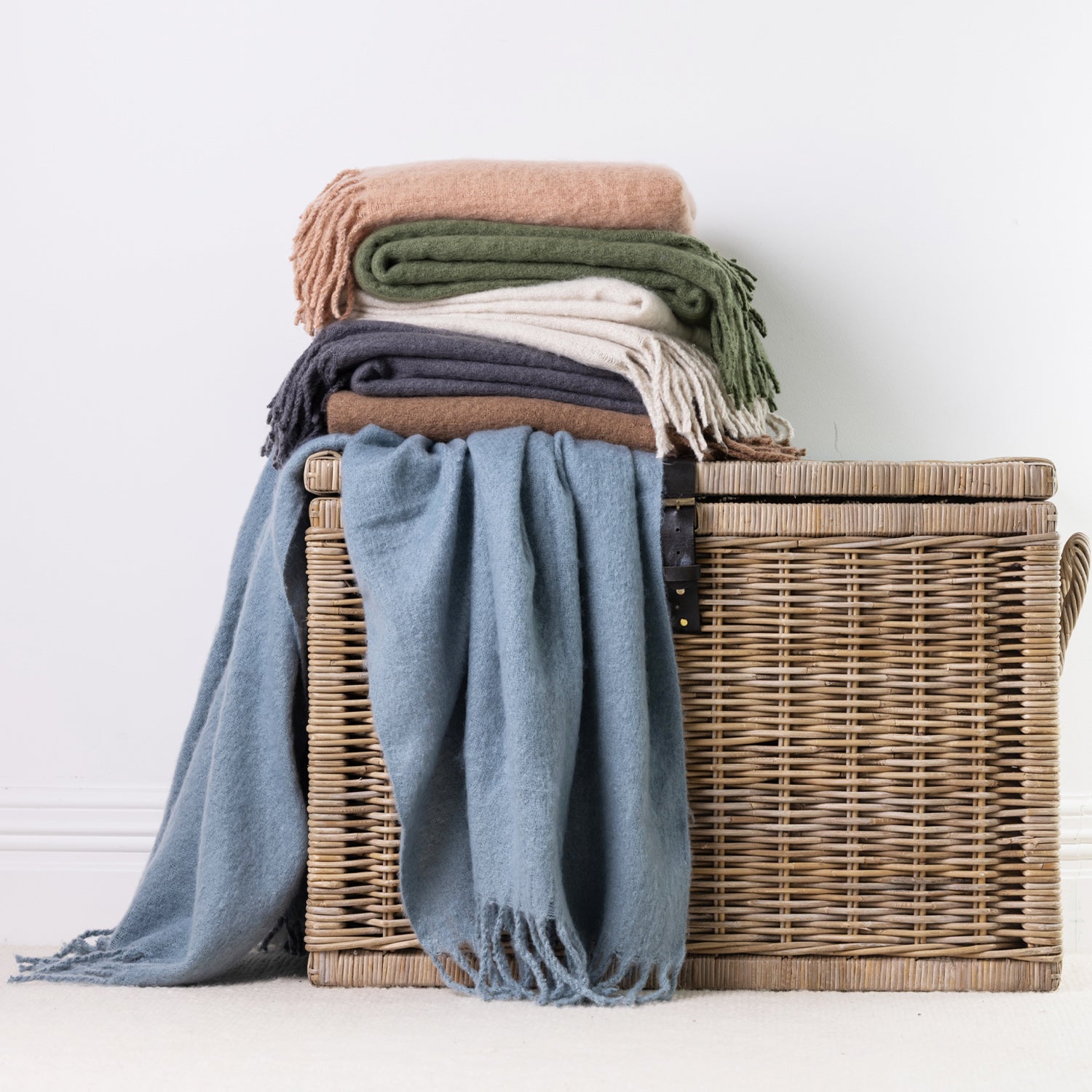 A stack of folded and draped faux mohair throws in blue, cocoa, green, natural, sand, and grey, arranged on and around a woven wicker storage basket against a plain white wall.
