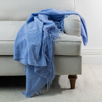 Blue: Solace Simone Throw in blue draped over the arm of a light grey sofa, featuring a textured woven finish and fringed edges.