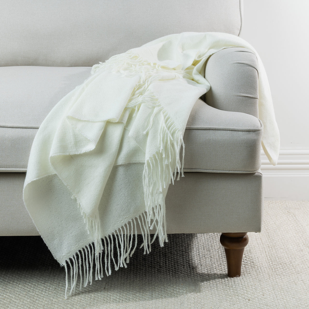 Ivory: Solace Simone Throw in ivory draped over a light grey sofa, featuring a soft woven texture and long fringed edges.