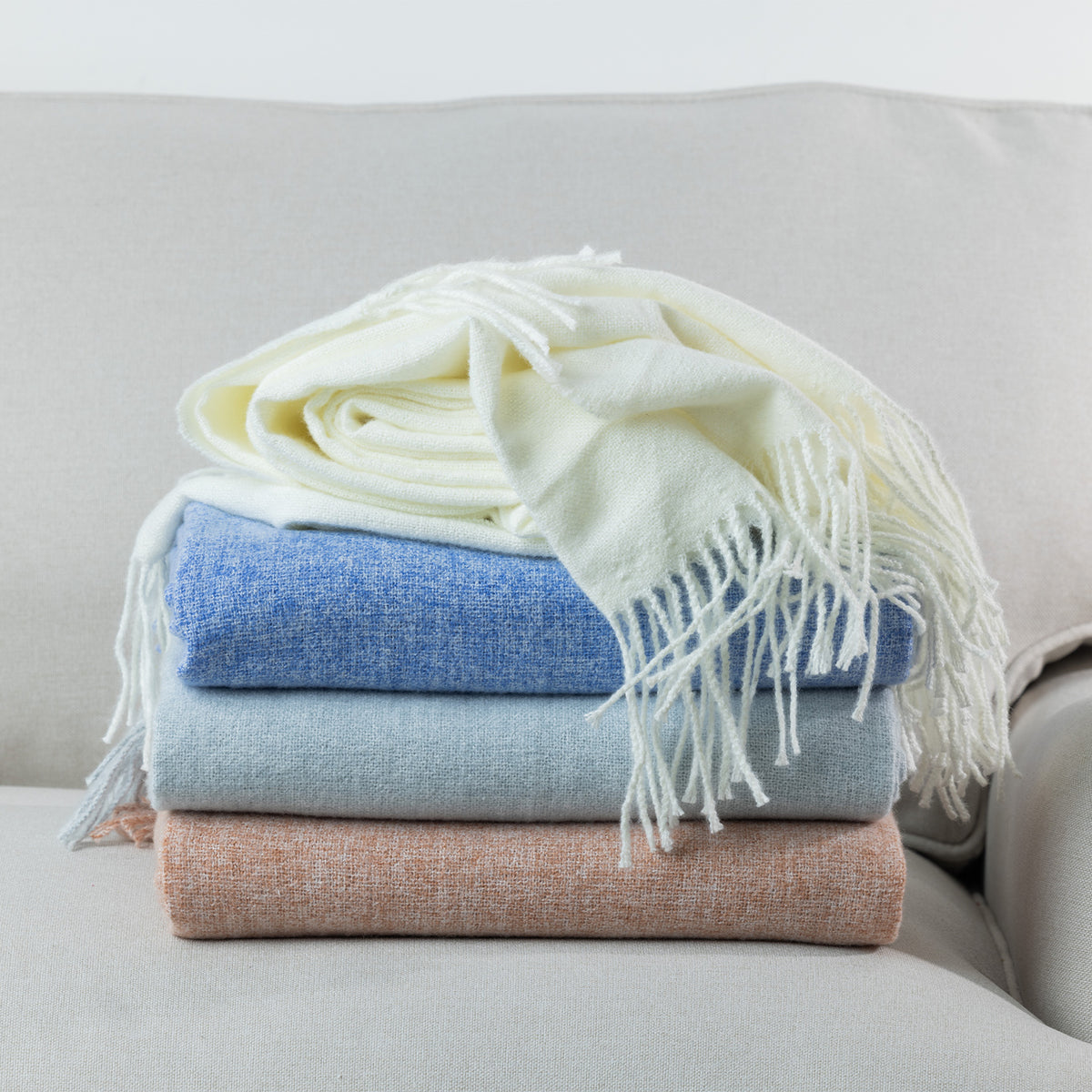 A stack of Solace Simone Throws in ivory, blue, silver and rust, showcasing their soft woven texture and fringed detailing.