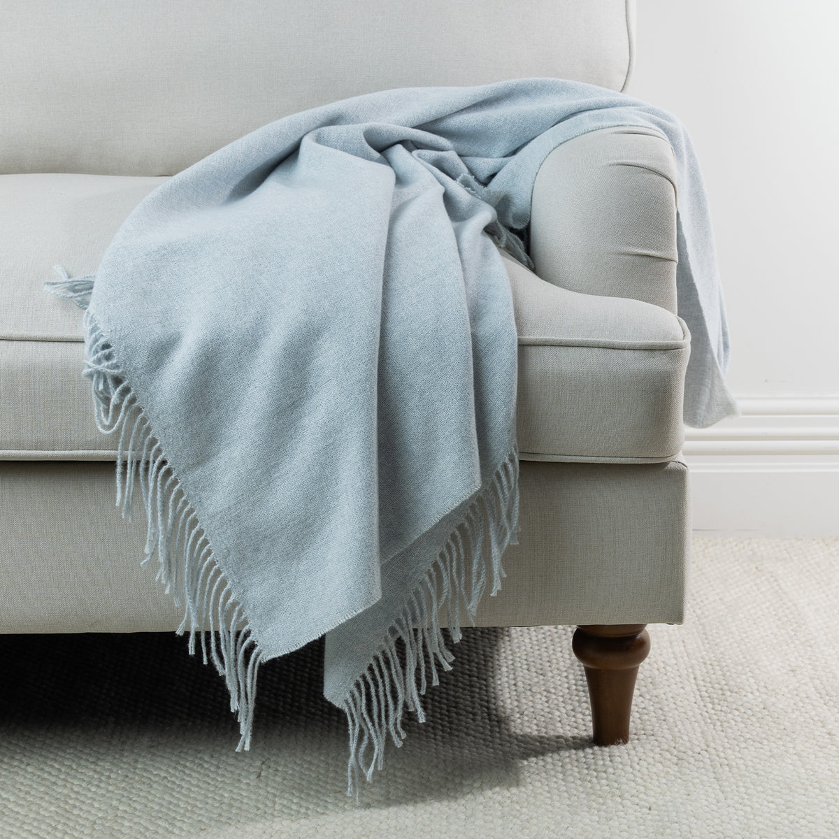 Silver: Solace Simone Throw in silver draped over a light grey sofa, showcasing its soft woven texture and fringed edges.