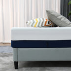Close-up side view of a white and navy mattress on a light grey upholstered bed base, styled with decorative cushions in a modern bedroom setting.