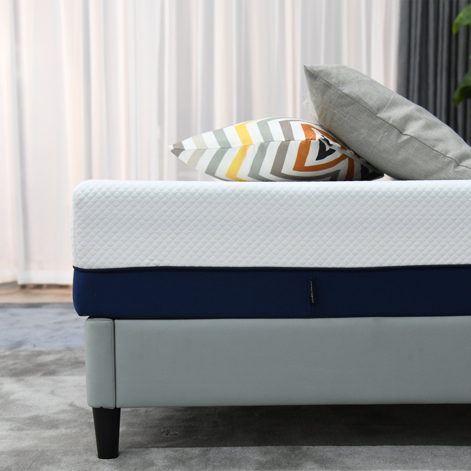 Close-up side view of a white and navy mattress on a light grey upholstered bed base, styled with decorative cushions in a modern bedroom setting.