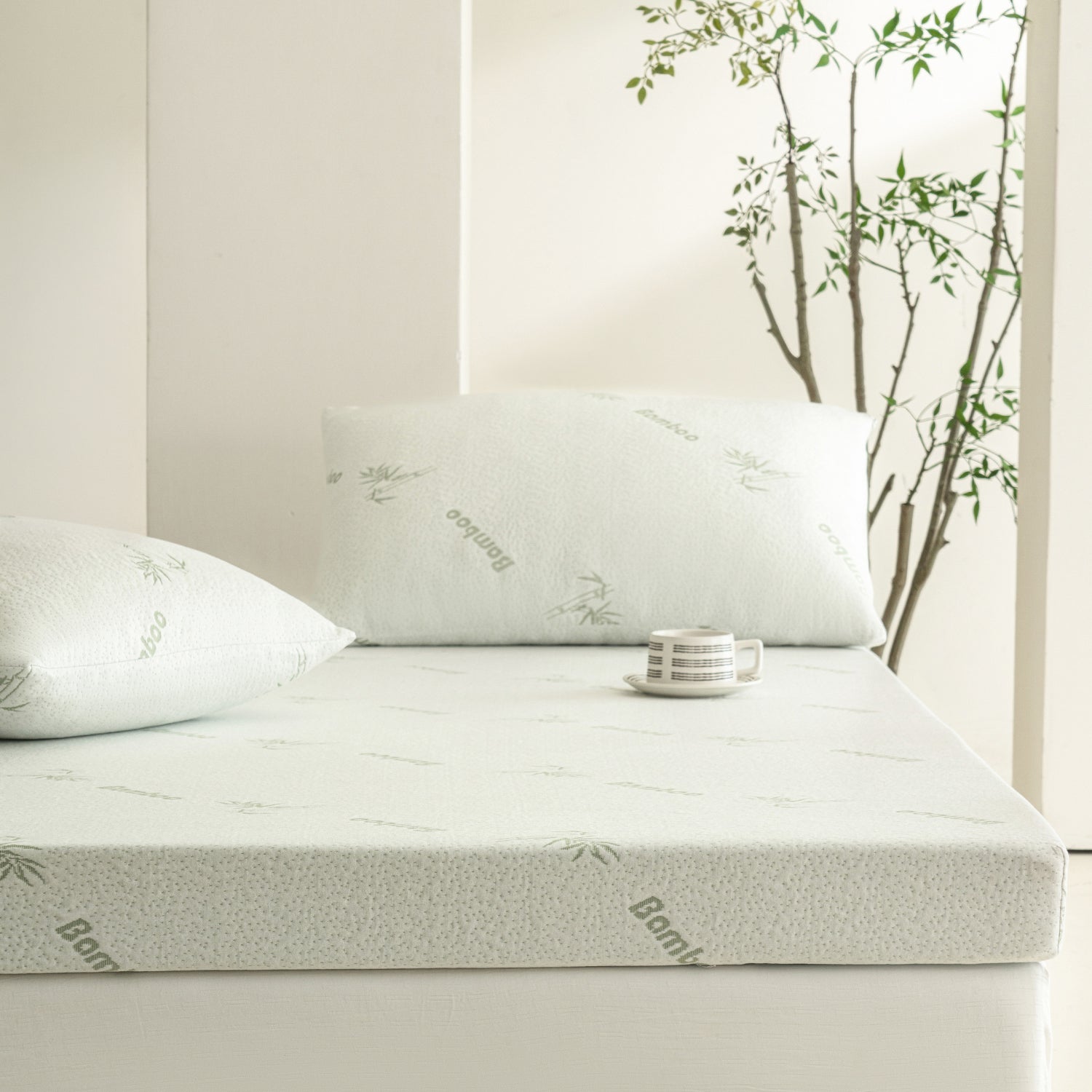 Bambou 10cm Memory Foam Mattress Topper displayed on a mattress with matching bamboo-patterned pillow, styled in a bright bedroom setting with a small cup and decorative indoor tree.