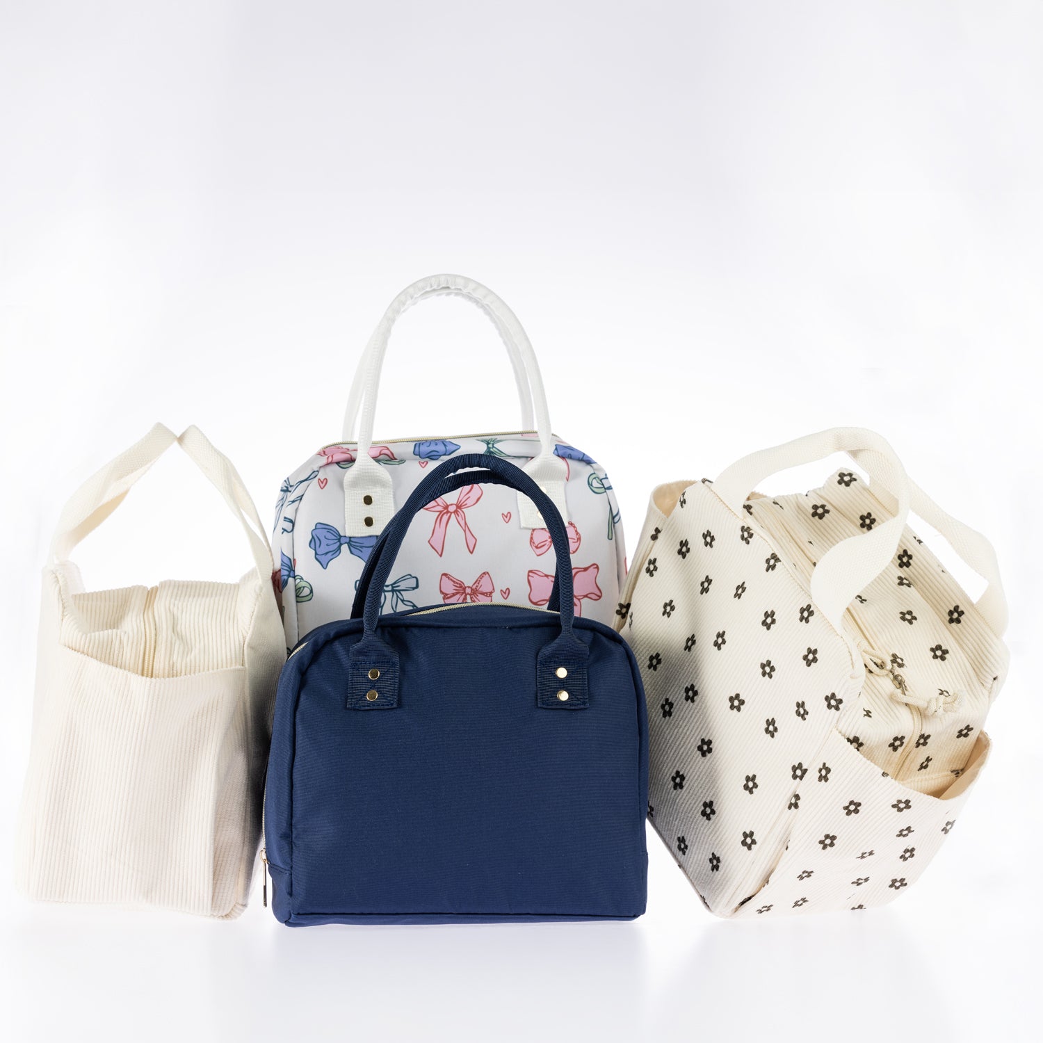 Assorted lunch totes in cream corduroy, navy, colourful bow print, and brown daisy print with carry handles on a white background.