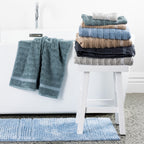 Home Co. Penny Self Stripe towels styled in a bathroom, with a stacked range of colours on a white stool, a seafoam towel draped over the bath, and a dusty blue bath mat on the floor.
