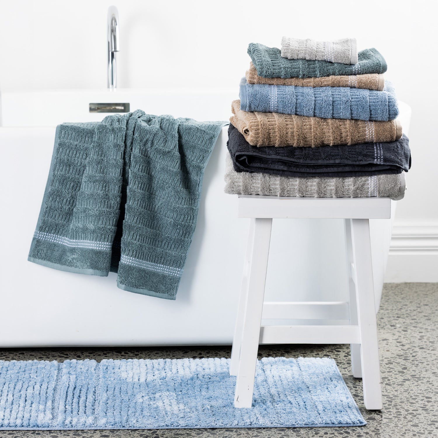 Home Co. Penny Self Stripe towels styled in a bathroom, with a stacked range of colours on a white stool, a seafoam towel draped over the bath, and a dusty blue bath mat on the floor.