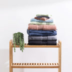 Assorted Fieldcrest Amelie plush towels neatly stacked on a wooden bathroom shelf beside a trailing green plant.
