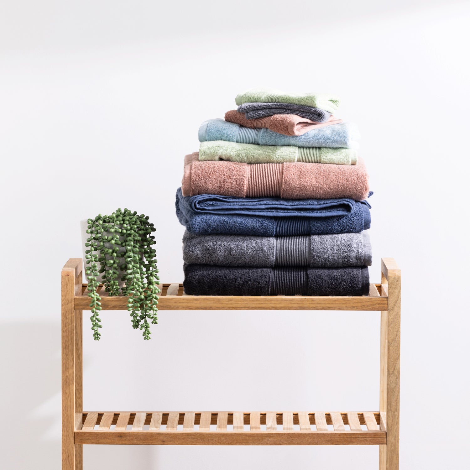 Assorted Fieldcrest Amelie plush towels neatly stacked on a wooden bathroom shelf beside a trailing green plant.