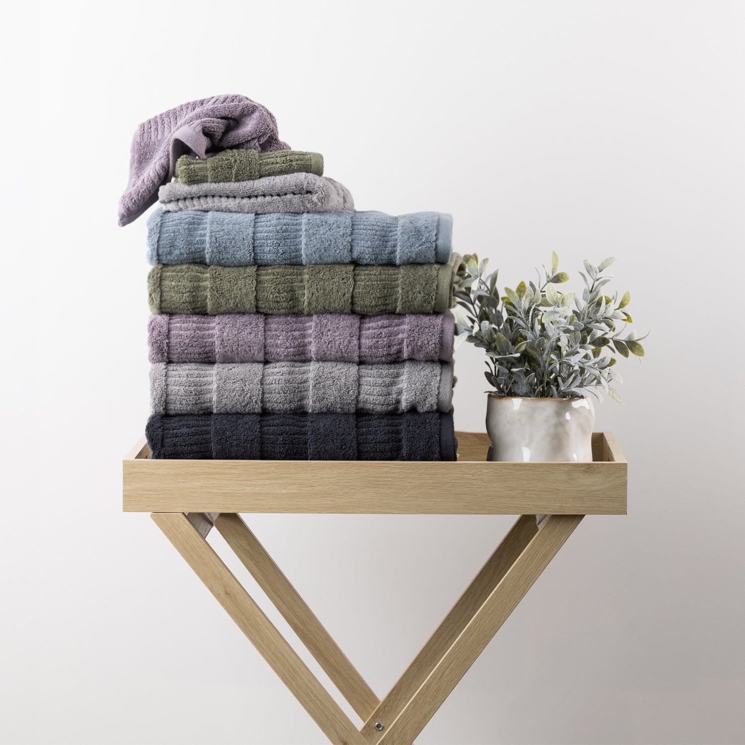 Assorted Design Republique Addison textured face cloths stacked with coordinating towels in faded blue, khaki, grape, silver and charcoal beside a decorative plant.