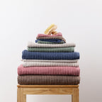 Coordinating Home Co. Lara Stripe towel range stacked in assorted colours on a wooden bathroom stool, with bath sheets, towels, hand towels and smaller towels layered together.