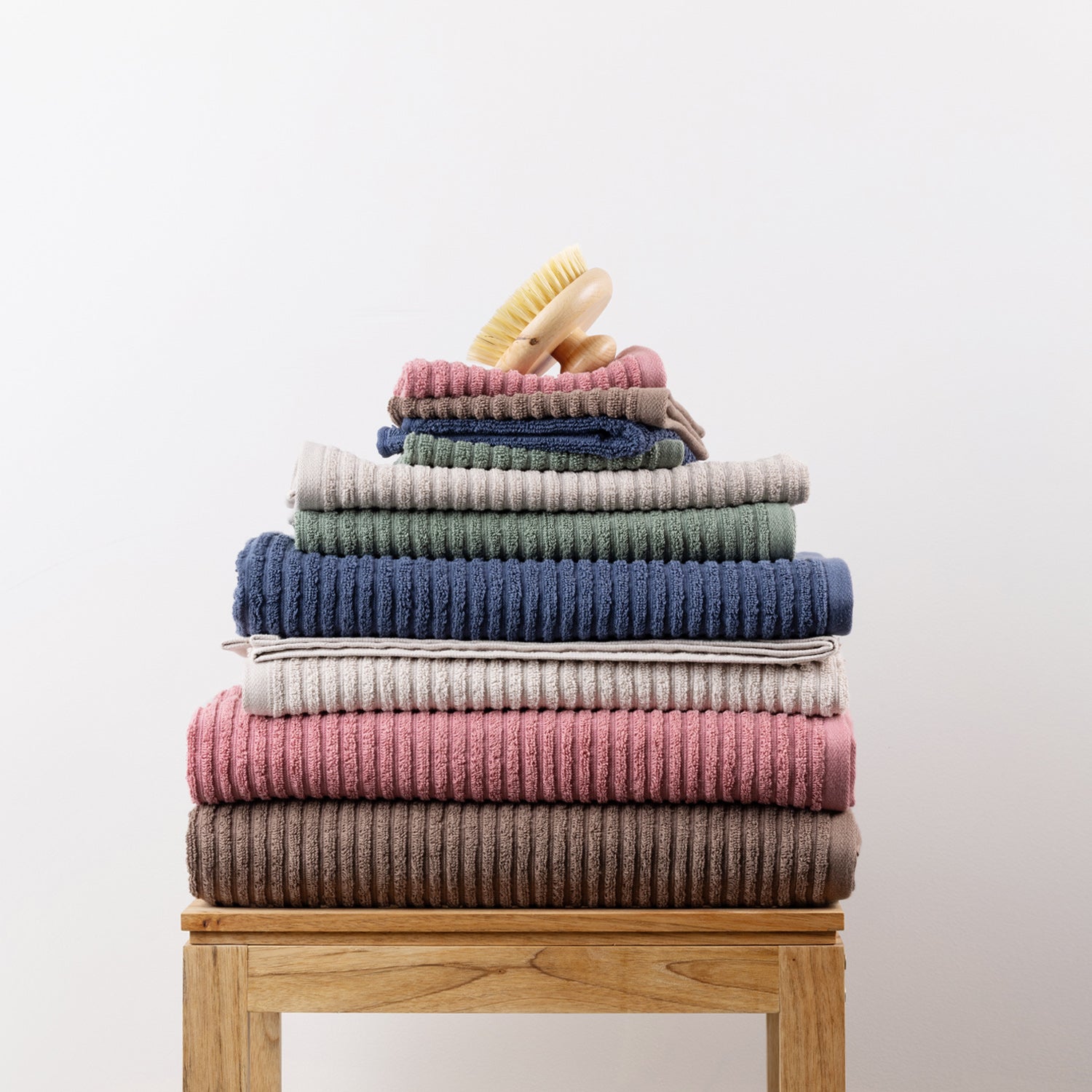Coordinating Home Co. Lara Stripe towel range stacked in assorted colours on a wooden bathroom stool, with bath sheets, towels, hand towels and smaller towels layered together.