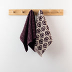 Two berry hand towels hanging from a wooden peg rack, featuring a deep berry textured towel and a coordinating cream and berry patterned towel with fringe.
