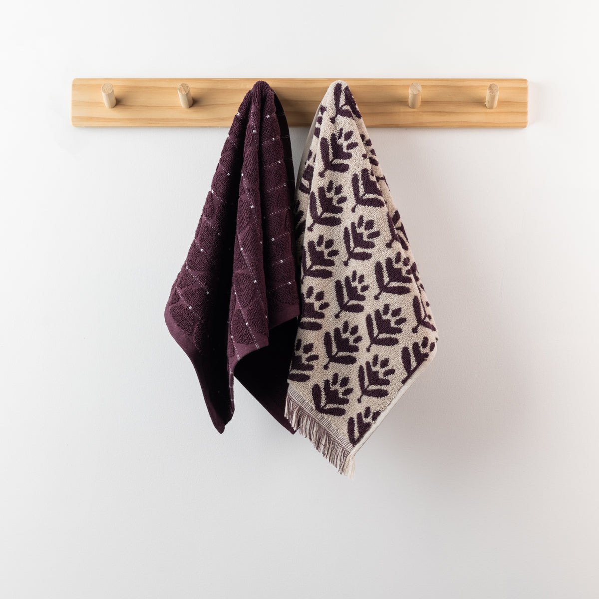 Two berry hand towels hanging from a wooden peg rack, featuring a deep berry textured towel and a coordinating cream and berry patterned towel with fringe.