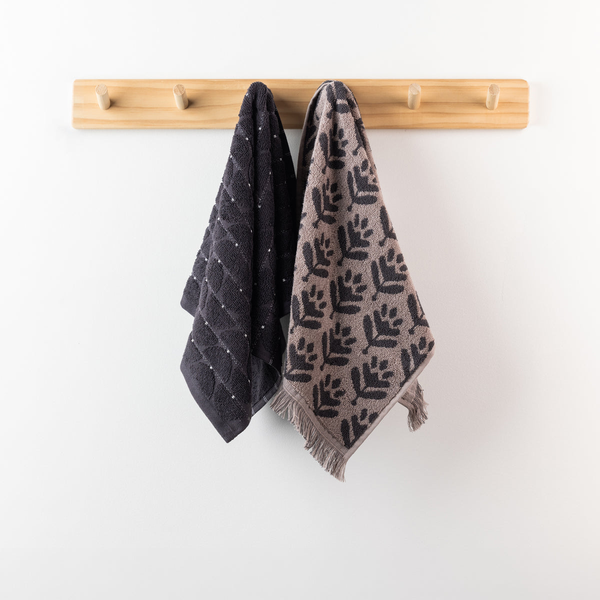 Two charcoal hand towels displayed on a wooden peg rack, including a dark charcoal textured towel and a coordinating taupe patterned towel with charcoal leaf motifs and fringe.