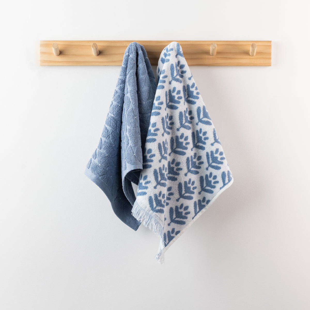 Two denim hand towels hanging from a wooden peg rack, featuring a solid textured denim blue towel and a coordinating white towel with blue leaf pattern and fringe.