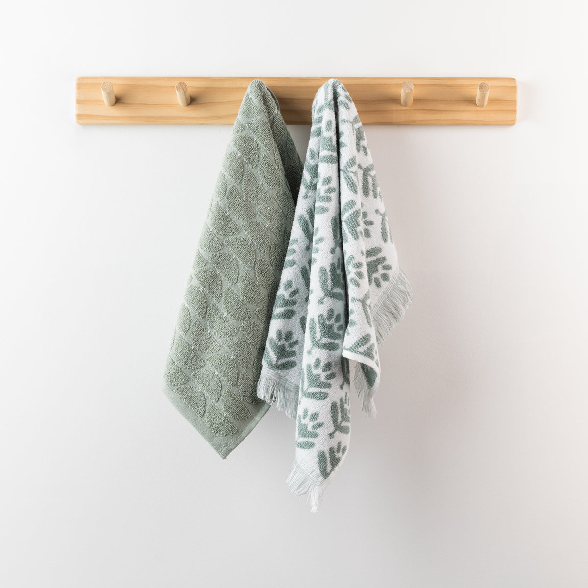 Two sea mist hand towels displayed on a wooden peg rack, including a solid textured towel and a coordinating white patterned towel with sea mist leaf motifs and fringe.