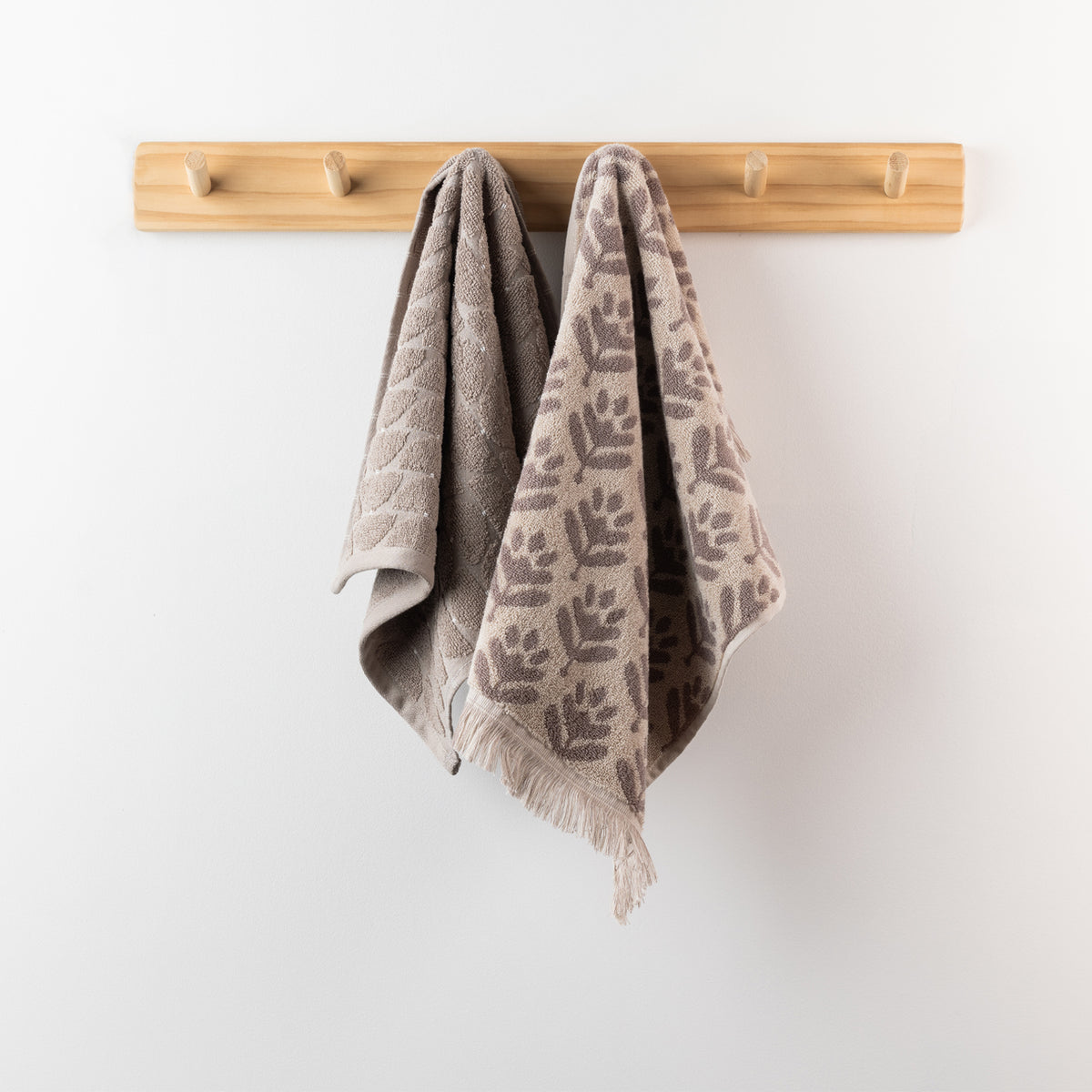 Two taupe hand towels hanging from a wooden peg rack, including a solid textured towel and a coordinating patterned towel with fringe detailing.