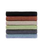 A neatly stacked collection of Home Co. Nevada Bath Towels featuring a textured cotton finish in black, rust, beige, sage green, charcoal and pale blue, displayed against a white background.