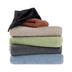 A stack of Home Co. Nevada Bath Towels featuring a textured cotton finish in black, rust, beige, sage green, charcoal and pale blue, with the top black towel partially unfolded to showcase its texture, displayed against a white background.
