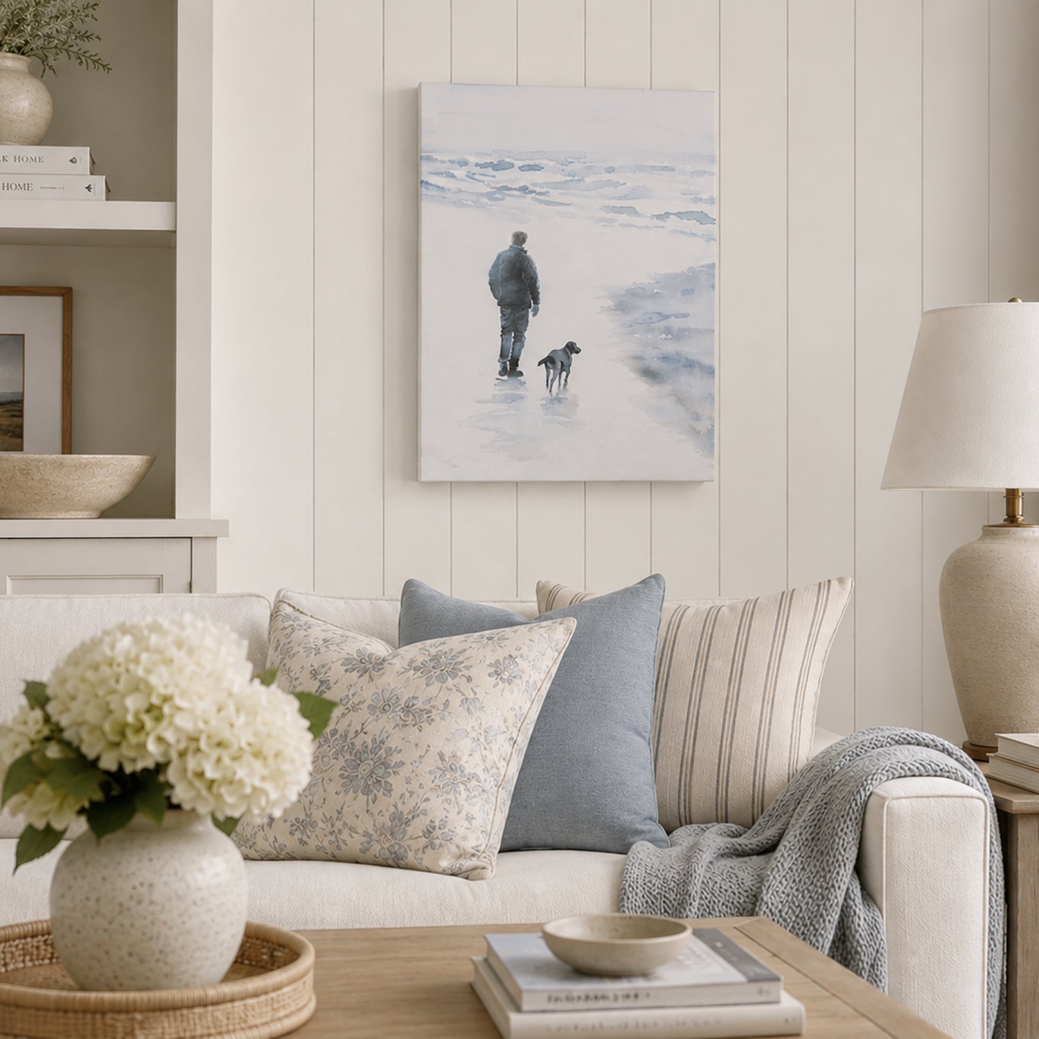 A soft coastal artwork depicting a person and their dog strolling along a tranquil shoreline, beautifully styled in a light, modern country living room with blue and neutral accents.