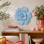 A large light blue flower wall decor piece with a textured, ribbed petal design, mounted on a beige wall above a wooden shelf styled with books, candles, and a vase of flowers.