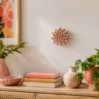 A pink sculptural dahlia flower wall decor piece mounted on a warm beige wall above a wooden console table styled with books, vases, and potted plants.