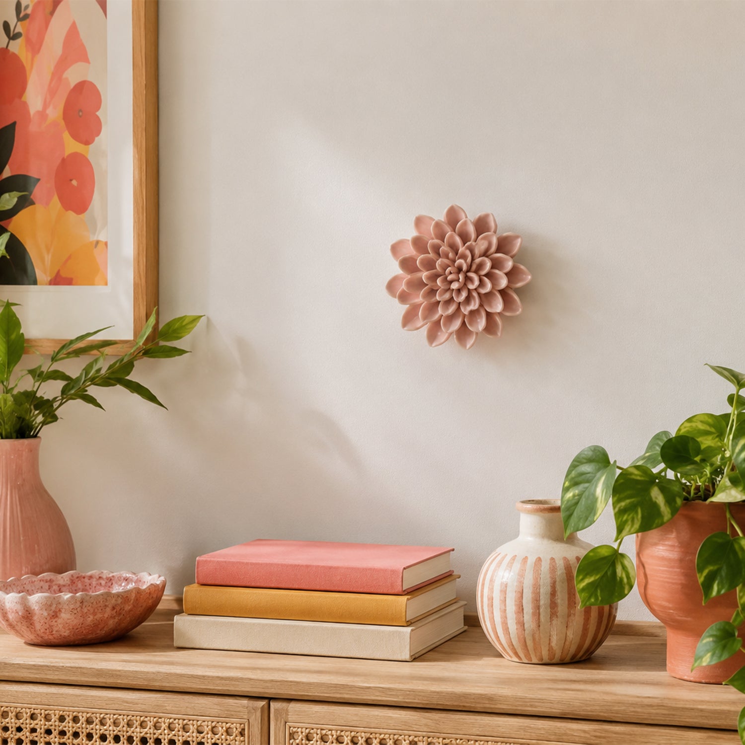 A pink sculptural dahlia flower wall decor piece mounted on a warm beige wall above a wooden console table styled with books, vases, and potted plants.